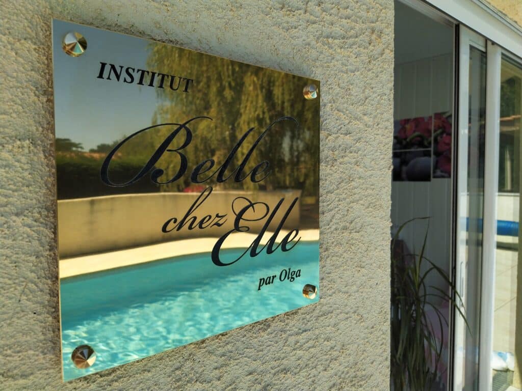 Plaque dorée gravée 'INSTITUT Belle chez Elle par Olga' fixée sur un mur crépi, reflétant une piscine ensoleillée.