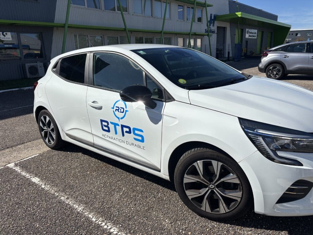 Véhicule BTPS Réparation Durable : Renault Clio de société Renault blanche siglée BTPS Réparation Durable garée devant des bureaux sous un soleil radieux.