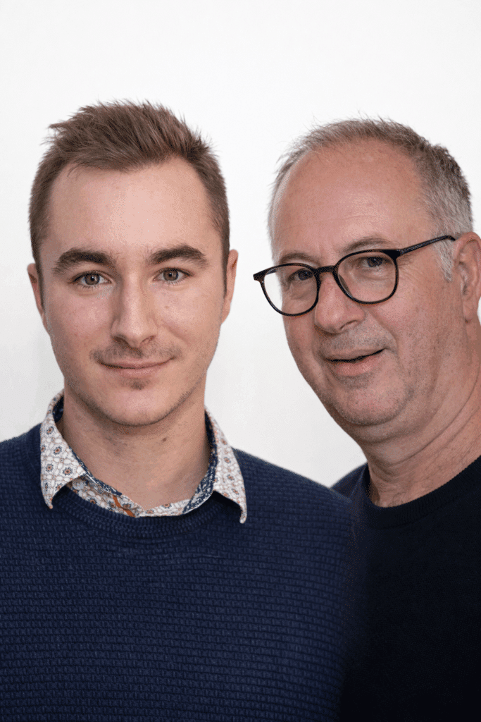 Duo d'hommes souriants : Portrait professionnel moderne Portrait professionnel de deux hommes souriants, l'un jeune en pull texturé bleu, l'autre plus âgé avec des lunettes, sur fond blanc.