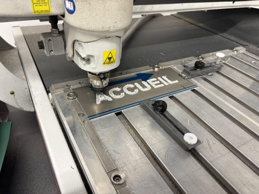 Machine gravant le mot ACCUEIL et une flèche bleue sur une plaque métallique industrielle.
